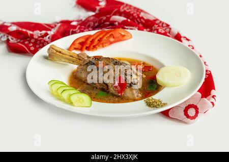 Geschmorte Lamb Shanks mit Sauce und Tomaten, Gurken, Rettich in einem Gericht isoliert auf bunten Tischtuch Draufsicht auf grauem Hintergrund Stockfoto