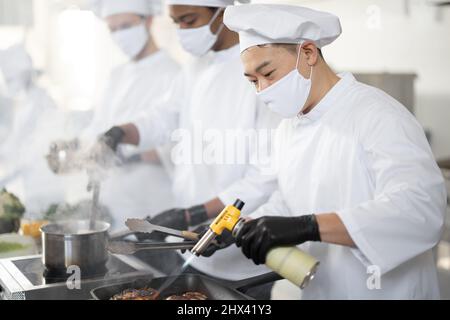 Multirassisches Team von Köchen kocht in weißer Uniform zusammen in der Küche. Asiatischer Koch, der Fleisch mit Brenner, lateinamerikanische und europäische Küche auf dem Hintergrund braten lässt. Köche tragen Gesichtsmasken und Schutzhandschuhe Stockfoto