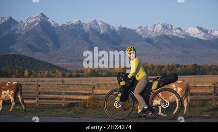 Die Frau reist auf gemischtem Terrain mit Bikepacking. Die Reise des Reisenden mit Fahrradtaschen. Sporttourismus Bikepacking, Fahrrad, Sportbekleidung in grün-schwarzen Farben. Schneebedeckte Berge. Stockfoto
