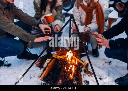 Eine Gruppe von Freunden, die sich am Lagerfeuer im Hinterhof treffen, Tee trinken und sich die Hände wärmen. Zwei glückliche Paare, die sich entspannen und die Wintersaison genießen, während sie sitzen Stockfoto