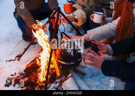 Eine Gruppe von Freunden, die sich am Lagerfeuer im Hinterhof treffen, Tee trinken und sich die Hände wärmen. Zwei glückliche Paare, die sich entspannen und die Wintersaison genießen, während sie sitzen Stockfoto