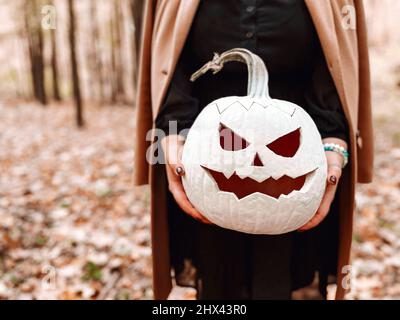 Nahaufnahme eines jungen blonden Weibchens, das beängstigend gefertigte weiße Kürbisse hält und isoliert über dem herbstlichen Waldhintergrund steht. Herbstferien, halloween d Stockfoto