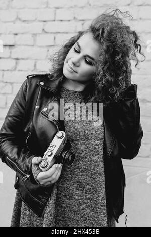 Monochromes Porträt einer jungen schönen fröhlichen Frau mit roten lockigen Haaren, die Bilder auf Film altmodische Kamera auf weißem Ziegelsteinwand Hintergrund Stockfoto