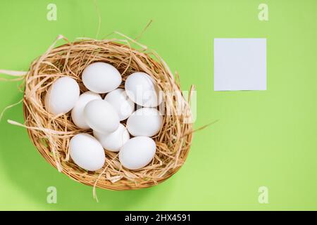 Weiße ostereier im gelben Korb mit Stroh auf grünem Hintergrund. Ansicht des Postkarten-Konzepts „Happy Easter“ von oben Stockfoto