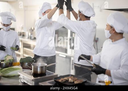 Multirassisches Team von Köchen, die sich fünf mit Händen geben, während sie in der professionellen Küche kochen. Konzept von Teamarbeit und Erfolg im Restaurantbetrieb. Köche tragen Gesichtsmasken und Schutzhandschuhe Stockfoto