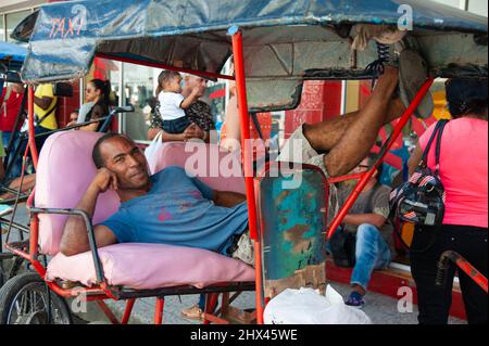 Fahrradtaxi-Fahrer entspannt sich auf dem Rücksitz seines Fahrradwagens, während die Menschen durch eine Straße in Cienfuegos, Kuba, laufen. Stockfoto