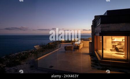 Blick von außen auf die Dämmerung von beleuchteten Fenstern und privatem Sitzbereich und Aussichtsplattform mit Meerblick und Sonnenuntergang. Clifftops, Dorset, Portland, United Stockfoto