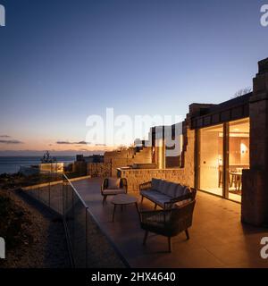 Blick von außen auf die Dämmerung von beleuchteten Fenstern und privatem Sitzbereich und Aussichtsplattform mit Meerblick und Sonnenuntergang. Clifftops, Dorset, Portland, United Stockfoto