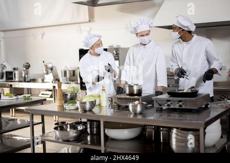 Multirassisches Team von Köchen in Uniform und Gesichtsmasken beim Kochen von Mahlzeiten für ein Restaurant in der Küche. Konzept der Teamarbeit im Restaurant während einer Pandemie. Lateinische, asiatische und europäische Jungs kochen zusammen Stockfoto