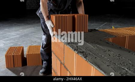 Industriearbeiter, der Ziegelsteine auf Zement legt, während er Wände baut. Nahaufnahme eines Mannes in Schutzhandschuhen, der schweren Ziegelstein-Block zu einem bildet Stockfoto