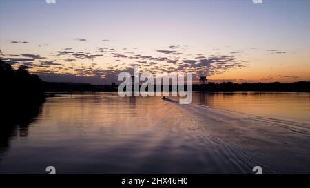 Spiegelung eines orangefarbenen Sonnenuntergangs im Wasser eines Stadtsees. Dunkle Silhouette von Bäumen, die am Ufer wachsen, und ein Motorboot, das einen welligen Weg in der Sonne verlässt Stockfoto