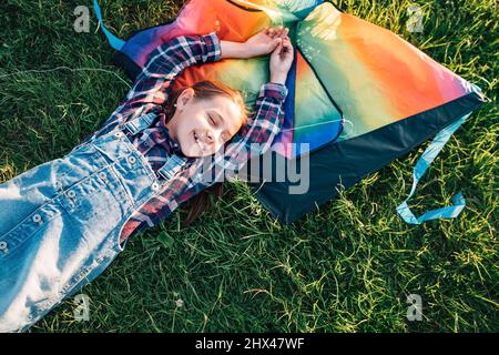 9YO lächelndes Mädchen, das auf dem Gras liegt, mit einem farbenfrohen Regenbogen-Drachen-Spielzeug. Glückliche Kindheitsmomente oder ein Konzept für die Zeit im Freien. Stockfoto