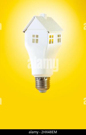 Schwebender Glühbirne in Form eines weißen Hauses, isoliert auf gelbem Hintergrund. Glühbirne in Hausform Stockfoto