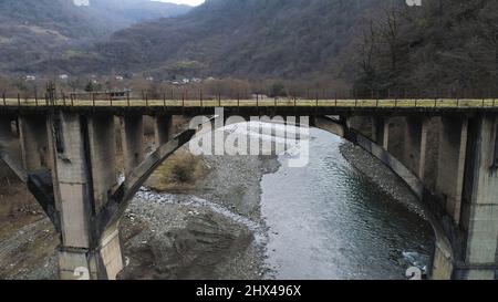 Fliegen Sie auf die alte Betonbrücke, die von grünem Moos bedeckt ist. Luftbild der ländlichen Landschaft mit einem kleinen Dorf in hügeliger Region. Stockfoto