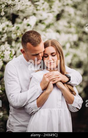 Junges, liebevolles Paar bei einem Spaziergang in einem frühlingsblühenden Apfelgarten. Glückliches Ehepaar genießen einander beim Spaziergang im Garten. Mann, der eine Frau hält Stockfoto