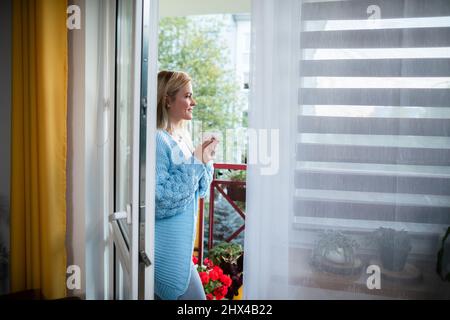 Ein Mädchen steht mit einer Tasse Tee in der Hand auf dem Balkon. Stockfoto