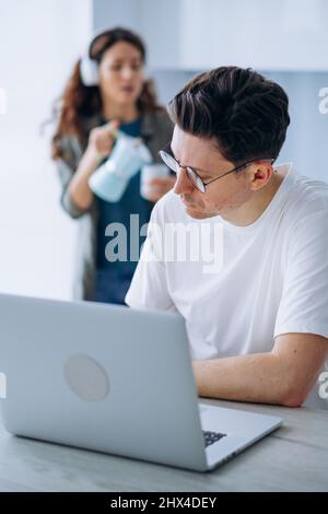 Lockig langhaarige Frau mit Kopfhörern lächelt und gießt Kaffee in den Becher für gestressten Ehemann, der am Laptop arbeitet und Arbeitsprobleme löst Stockfoto