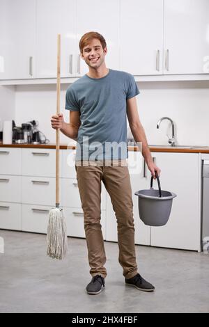 Bereit, die Hausarbeiten zu erledigen. In voller Länge ein hübscher Mann, der in der Küche mit einem Wischmopp und einem Eimer steht. Stockfoto