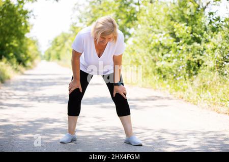 Fitness-blonde Frau müde nach dem Joggen im Wald auf der Straße ruht mit ihren Armen auf den Knien, trägt Sportkleidung und Smart Watch an Stockfoto
