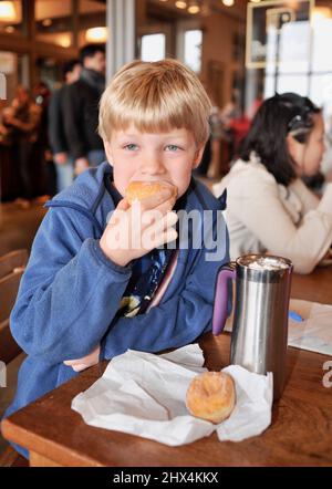 Kleines Kind, das ein bisschen in einen leckeren frischen Donut steckt, der am Tisch im Ferry Building Marketplace, San Francisco, Kalifornien, USA, sitzt Stockfoto
