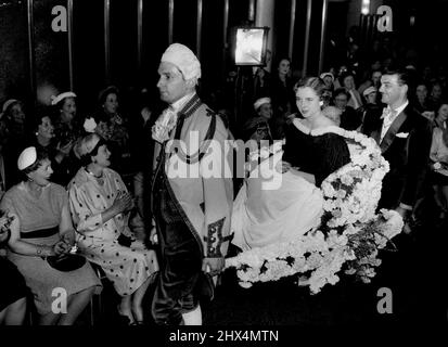 Chairborne Style -- Raymond, der Friseur von „Teazle-Weazie“ im Fernsehen, hilft einem unverschämtem, den blumengeschmückten Sedan-Stuhl mit dem Modell Claudette zu tragen, der bei der Eröffnung seines neuen Salons in Cardiff, Südwales, eine seiner Herbstmoden zeigt. Claudettes Coiffure heißt „Cresta“. 03. September 1954. Stockfoto