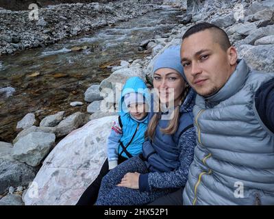 Touristen ruhen sich nach der Wanderung aus und machen Selfie am Telefon. Fotos vom Telefon während der Wanderung. Familienreisen. Menschen Umwelt durch Berge, Flüsse, Bäche. Papa Mutter und Tochter Sport reisen Familie. Stockfoto