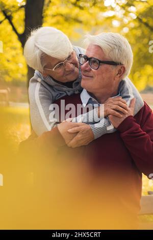Vertikale Aufnahme von zwei älteren grauhaarigen kaukasischen Menschen, die sich umarmen und Zeit im Freien verbringen, um den farbenfrohen Herbst zu genießen. Hochwertige Fotos Stockfoto