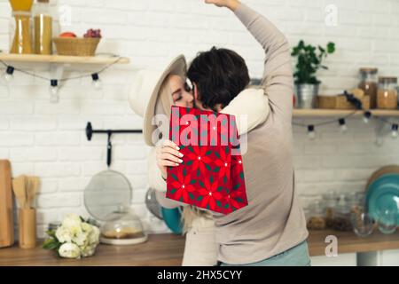 Der Ehemann mit Brunette mittleren Alters überrascht seine geliebte, wunderschöne blonde Frau mit einem Geschenk zum Geburtstag, während er in der modernen Küche steht. Hochwertige Fotos Stockfoto