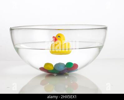 Gummiente schwimmt auf dem Wasser und bunte Tonkugeln am Boden der Schüssel Stockfoto