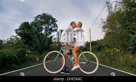 Radtour. Liebespaar auf blauem Fahrrad in weißer Kleidung auf Waldstraße. Gerade verheiratete Frau und Mann küssen, umarmen, stehen auf dem Fahrrad. Hochzeit, Flitterwochen. Cycling Cycle Fix. Asien Thailand Reiten Tourismus. Stockfoto