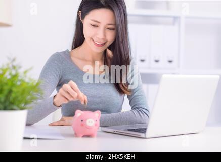 Junge Frau, die im Heimbüro Münzen in die Sparschwein legt Stockfoto