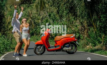 Zwei kaukasische Touristen Mann tanzen in der Nähe von Roller. Liebespaar auf rotem Motorrad in weißer Kleidung, Sonnenbrille auf Waldweg-Tour. Dancing Road. Motorradmiete. Asien Thailand Reiten Tourismus. Stockfoto