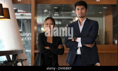 Selbstbewusste und erfolgreiche asiatische junge Geschäftsfrauen und Geschäftsleute in formellen Anzügen stehen mit gefalteten Armen in ihrem Büro. Stockfoto