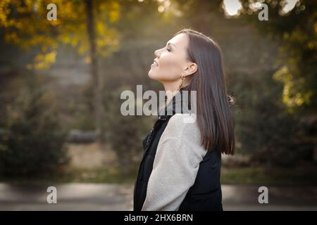 Profil einer jungen, ernsthaft nachdenklichen Frau mit Herbst im Park im Freien in einer warmen beigen Jacke und einer schwarzen ärmellosen Jacke. Hochwertige Fotos Stockfoto