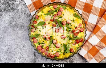 Wurst Frittata mit Käse, Rucola, Kartoffeln in einer Glasbackform auf dunkelgrauem Hintergrund. Draufsicht, flach liegend Stockfoto