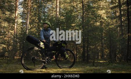 Die Frau reist auf gemischtem Terrain mit Bikepacking im Freien. Die Reise des Reisenden mit Fahrradtaschen. Stylische Bikepacking-, Bike-, Sportbekleidung in grün-schwarzen Farben. Magischer Waldpark. Stockfoto