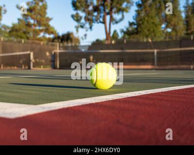 Gelber Tennisball auf grünem Platz mit schwarzem Netz und baumlichem Hintergrund. Rote Umschließfläche. Stockfoto