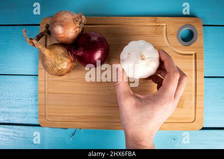 Knoblauch in der Hand einer Frau. Zwiebeln auf einem Bambusschneidebrett. Stockfoto