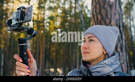 Die professionelle Videografin, die im Winter Smartphone auf einem 3-Achsen-Gimbal-Stabilisiergerät hält. Pro Equipment hilft, qualitativ hochwertige Videos auf dem Telefon zu erstellen. Kameramann. Zeitlupe Stockfoto