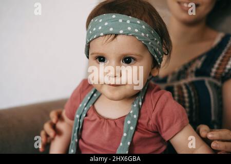 Porträt eines kleinen Mädchens, das zu Hause in den Armen ihrer Mutter ein Stirnband mit Punktmuster trägt. Familienbetreuung. Familienkonzept. Positive Person. Babypflege Stockfoto