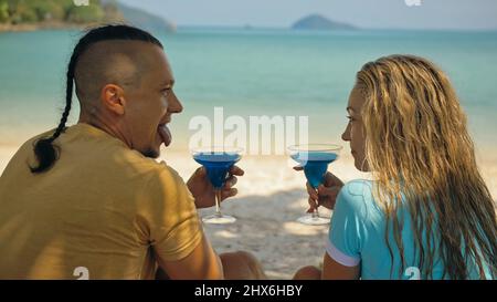 Das Liebespaar hält ein Glas blauen curacao Cocktail auf dem Meer. Mann mit Zopf und blonde Frau trinken Alkohol am Sandstrand im Schatten des azurblauen Ozeans einer tropischen Insel. Sommerferienkonzept. Stockfoto