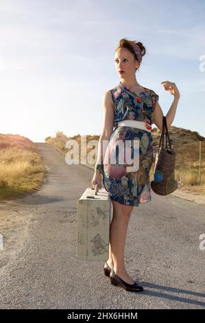 Frau mittleren Alters mit einem Vintage Pin-up-Stil mit einem Retro-Koffer und einer Handtasche. Haare mit einer Schleife gebunden, bereit für eine Reise zu gehen. Voyage Concept Stockfoto