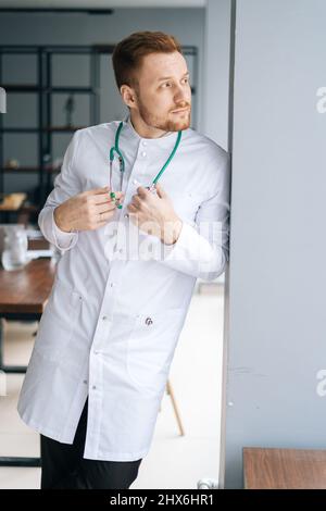 Vertikales Porträt eines nachdenklichen männlichen Arztes in weißer medizinischer Uniform, stehend mit Stethoskop in der Nähe des Fensters im Krankenhausbüro, wegblickend. Stockfoto