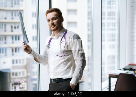 Porträt eines hübschen männlichen Arztes in weißer Uniform, der Röntgenaufnahme der Brust in der Nähe des Fensters im Krankenhausbüro untersucht, Stockfoto