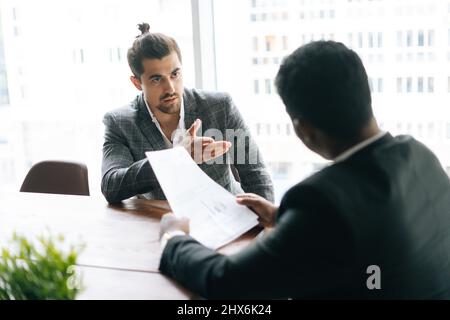 Blick von hinten auf zwei fokussierte, multiethnische Mitarbeiter, die am Schreibtisch in einem modernen Bürozimmer sitzen und über Dokumente diskutieren. Stockfoto