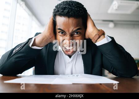 Nahaufnahme aus dem unteren Winkel Gesicht des beunruhigten empörten verwirrten afroamerikanischen Geschäftsmanns, der den Kopf hält und schreit. Stockfoto