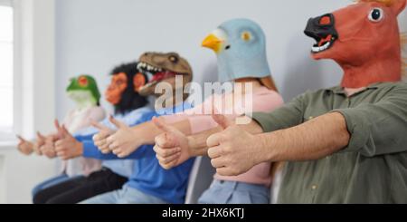 Verrückte exzentrische junge Menschen in trendigen Tiermasken sitzen in Reihe und zeigen Daumen nach oben. Stockfoto