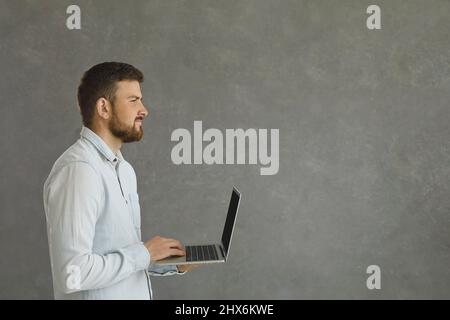 Porträt eines jungen und selbstbewussten Mannes, der auf einem grauen Hintergrund mit einem Laptop in den Händen steht. Stockfoto