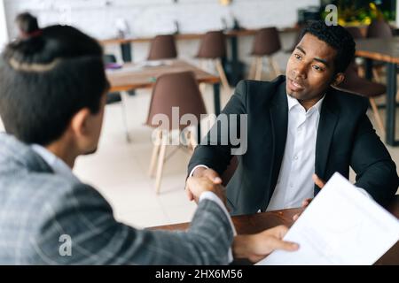 Nahaufnahme von zwei zufriedenen, multirassischen Geschäftsleuten, die nach erfolgreichen Verhandlungen die Hände über den Schreibtisch schüttelten und den Deal abschließen. Stockfoto