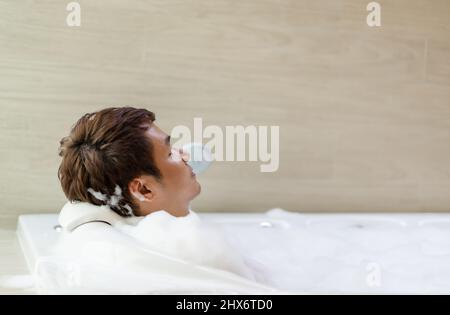 Junger Mann, der sich in der Badewanne mit geschlossenen Augen im Badezimmer entspannen kann Stockfoto